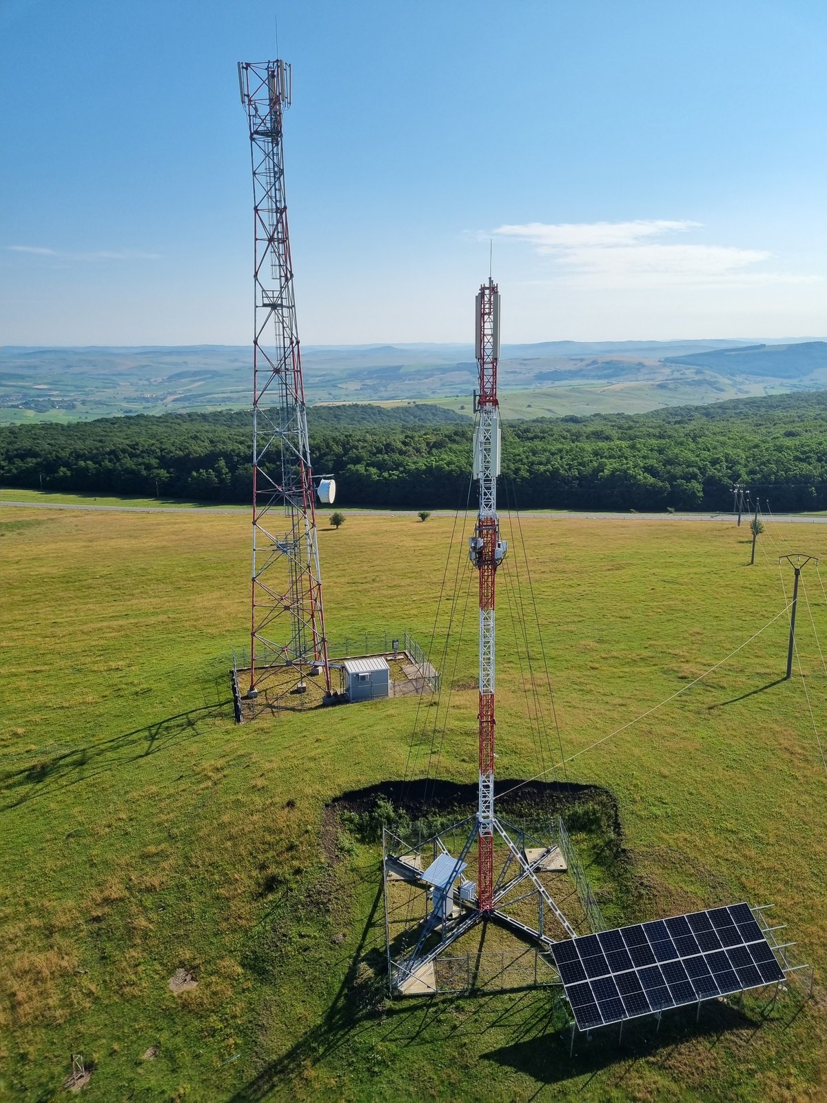 Site radio pe un deal verde - piloni cu antene, panou solar și pădure în fundal