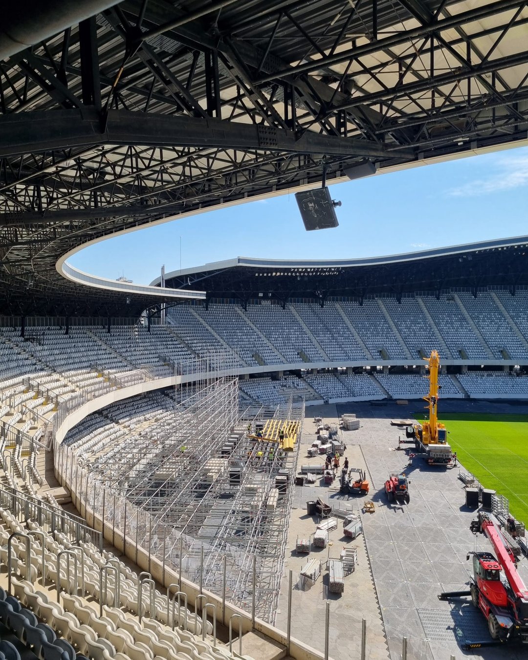 Vedere din tribună spre gazonul protejat al Cluj Arena, cu macarale și utilaje de ridicat în fereastra de instalare