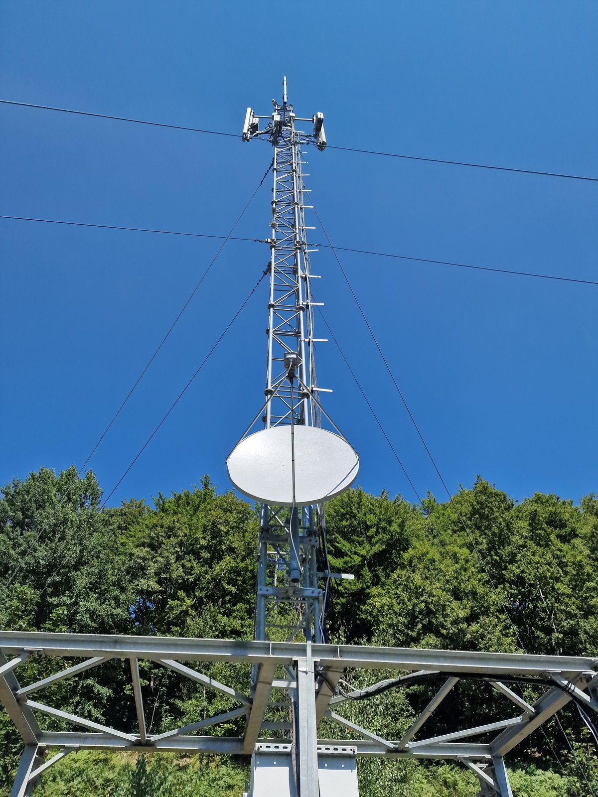 Pilon zăbrelit ancorat într-un site montan împădurit, cu antena VSAT montată la baza pilonului